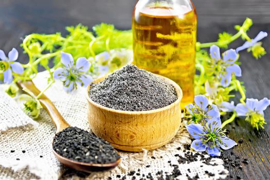 Flour of black caraway in a bowl, seeds in a spoon burlap napkin, oil in bottle and twigs Nigella sativa with blue flowers and leaves on dark wooden board background