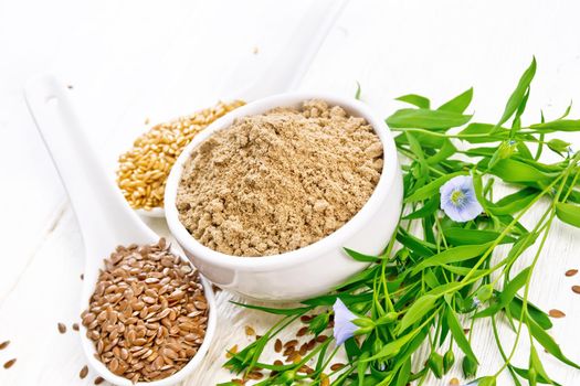 Flaxseed flour in a bowl, white and brown linen seeds in two spoons and on table, leaves and blue flaxen flowers on wooden board background