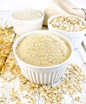 Meal oat, bran and oat flakes in three white bowls, oat stalks, towel on a background of wooden boards