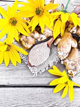 Jerusalem artichoke flour in a spoon on a burlap with yellow flowers and vegetables on wooden board background from above