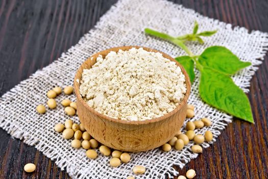 Soy flour in the bowl, soybeans on burlap, green leaf against the background of dark wooden boards
