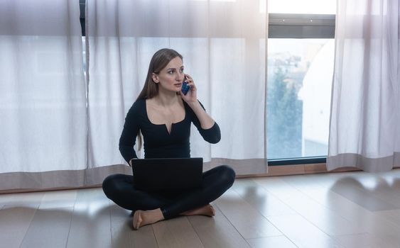 Self-quarantined woman with phone and laptop sitting on floor talking on the phone