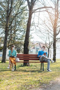 Woman and man with face mask in social distancing flirting sitting on a park bench
