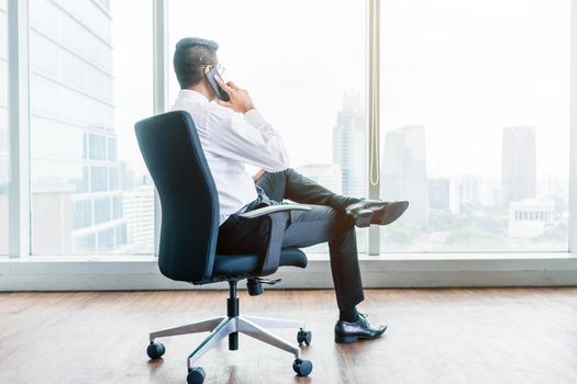 Rear view of businessman talking on phone while sitting down in an office with panoramic city view