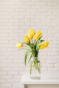 Bouquet of yellow tulips in a glass vase on a white brick wall background