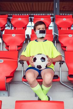 Lonely boy with football in his hand in soccer stadium watching a game wearing a mask