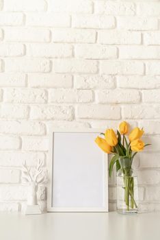 Bouquet of yellow tulips in a glass vase, white candle and blank photo frame on a white brick wall background. Mock up design