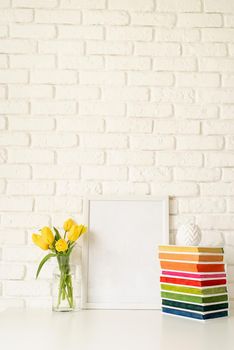 Bouquet of yellow tulips in a glass vase, pile of colorful books and blank photo frame on a white brick wall background. Mock up design