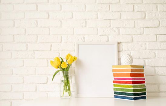 Bouquet of yellow tulips in a glass vase, pile of colorful books and blank photo frame on a white brick wall background. Mock up design