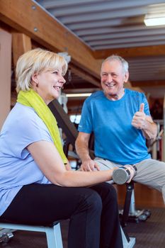 Senior woman in the gym exercising with dumbbells for fitness while her husband fires her up