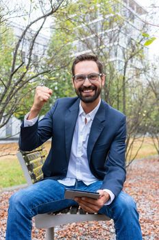 Portrait of successful businessman sitting on park bench holding digital tablet clenching his fist