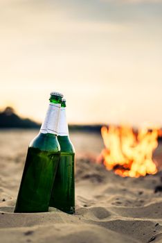 Two green beer bottles on sand in front of campfire at beach