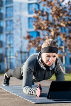 High-angle view of a fit young woman watching a motivational video on tablet PC, while exercising the forearm plank position on a mat outdoors in the park