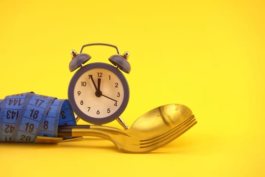 Golden cutlery wrapped with measuring tape near alarm clock on yellow background, weight loss or diet concept with copy space