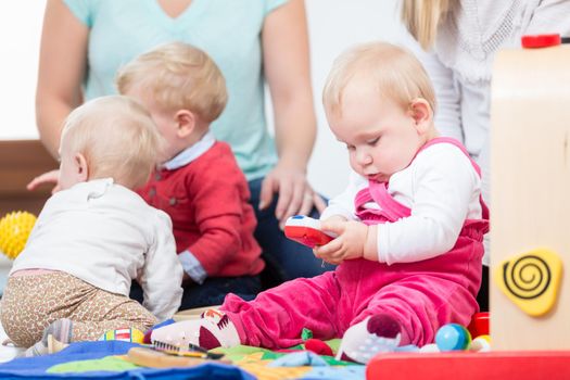 Three dedicated and happy young mothers sitting together on the floor, while watching their babies playing with safe multicolored toys at a modern playground for infants