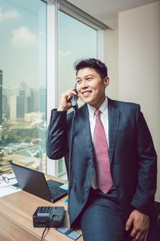 Portrait of happy businessman sitting on desk talking on telephone in the office
