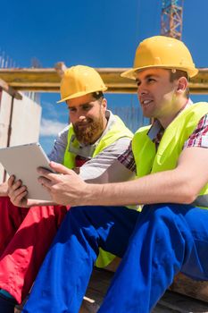Side view of two workers, reading online information or watching a video on a tablet PC during break at work on the construction site of a contemporary building