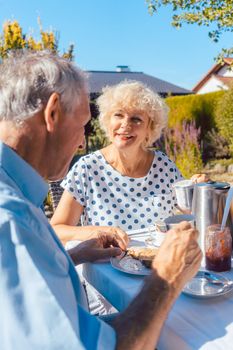 Happy elderly woman and man eating breakfast sitting in their garden outdoors in summer, eating bread rolls and drinking coffee