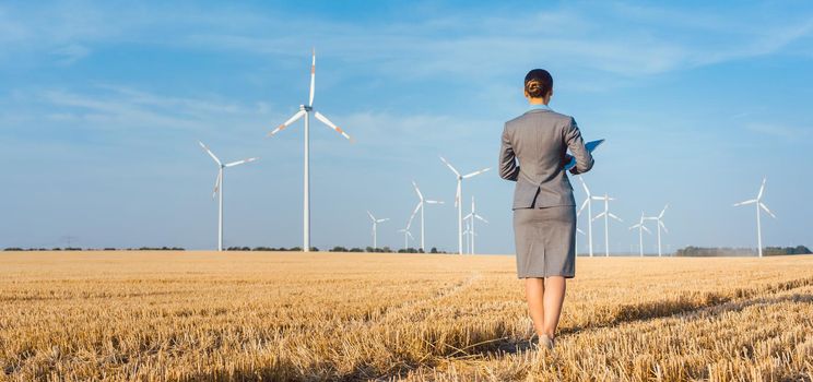 Investor in green energy looking at her wind turbines standing with suit on field