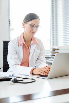 Doctor doing paperwork on computer sitting on desk in her office