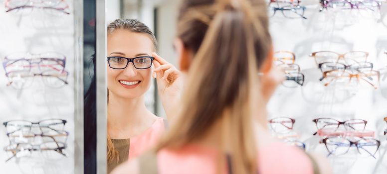Young woman trying fashionable glasses in optometrist store looking at herself in the mirror