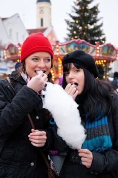 Two women on Christmas market eating cotton candy in front of a booth, it is cold