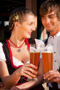 Couple in traditional Bavarian Tracht - Dirndl and Lederhosen - in front of a beer tent at the Oktoberfest or in a beer garden enjoying a glass of tasty wheat beer