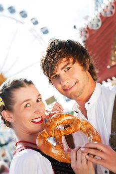 Young and beautiful couple in traditional Bavarian Tracht - Dirndl and Lederhosen - embracing each other on a fair like a Dult or the Oktoberfest eating a pretzel