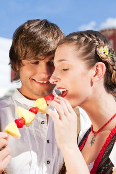 Young and beautiful couple in traditional Bavarian Tracht - Dirndl and Lederhosen - embracing each other on a fair like a Dult or the Oktoberfest eating traditional fruits