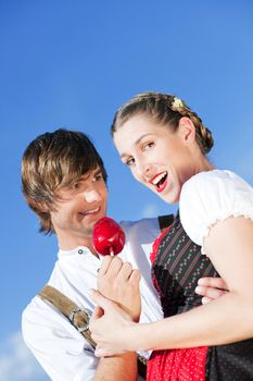 Young and beautiful couple in traditional Bavarian Tracht - Dirndl and Lederhosen - embracing each other on a fair like a Dult or the Oktoberfest eating traditional sugar apple