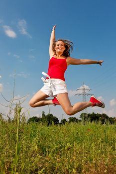 Woman in red t-shirt jumping high on a summer meadow under a blue sky, in the background a power pole is to be seen