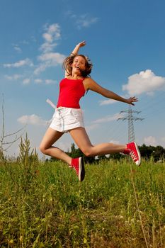 Woman in red t-shirt jumping high on a summer meadow under a blue sky, in the background a power pole is to be seen