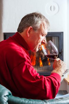 Mature man drinking a glass of red wine in front of his fireplace