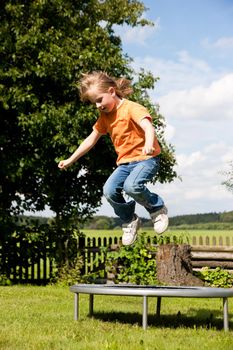 Little girl on a trampoline in a garden, in the background a fence and some landscape