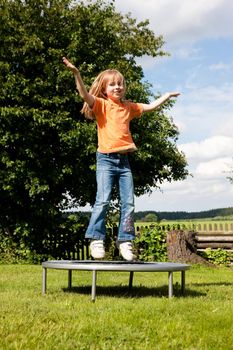 Little girl on a trampoline in a garden, in the background a fence and some landscape