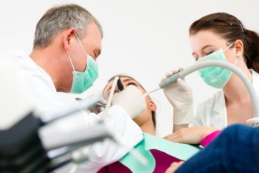 Female patient with dentist and assistant in a dental treatment, wearing masks and gloves, Dentist and assistant bowing over her using sucker and drill