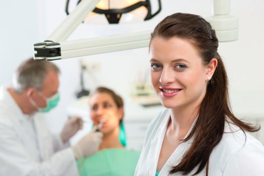 Dentists in her surgery looking at the viewer, in the background her colleague is giving a female patient a treatment