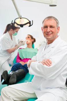 Dentists in his surgery looking at the viewer, in the background his assistant is giving a female patient a treatment