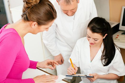 Patient in reception area of office of doctor or dentist, handing her health insurance card over the counter to the nurse who is writing things on a clipboard, the doctor standing in the background 