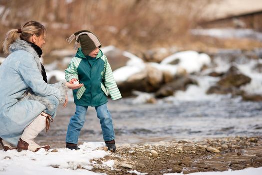 Family - mother and son to be seen - on a walk along a riverbank in winter; the child is throwing a snowball