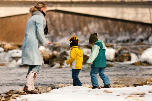 Family - mother and sons to be seen - on a walk along a riverbank in winter; the child is throwing a snowball