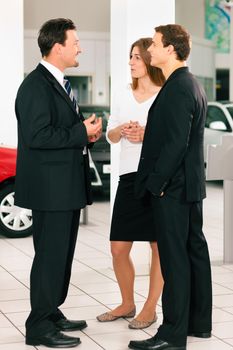 Sales situation in a car dealership, the dealer is talking to a young couple, there are cars standing in the background