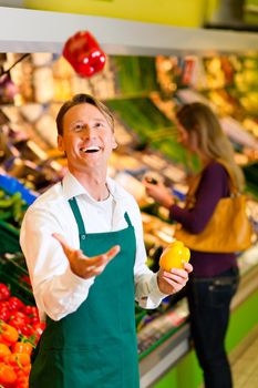 Shop assistant in a supermarket at the vegetable shelf; he is tossing a bell pepper