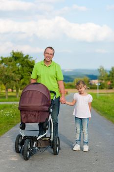 Father with child and a baby lying in a baby buggy walking down a path outdoors