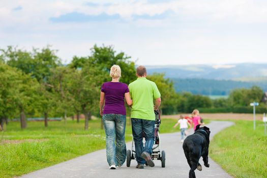 Family with three children (one baby lying in a baby buggy) walking down a path outdoors, two kids are running ahead, there is also a dog