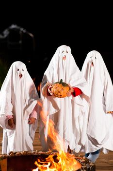 Three very, very scary spooks - kids dressed as ghosts - on Halloween or for carnival or a costume party in front of a fire; FOCUS IS ON PUMPKIN