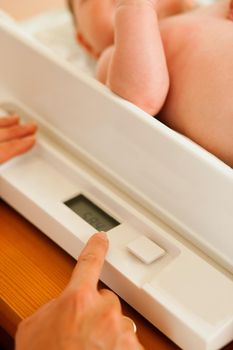 Baby on a weight scale, her mother or a doctor is checking health and development of the newborn