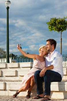 Couple having a city break in summer sitting on a brick wall in the sunlight, making pictures