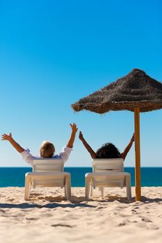 Couple sitting in sun chairs under an parasol sunshade on a beach stretching arms, feeling free