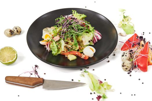 Top view of modern black plate with appezing vegetables salad on white background. Concept of process cooking with knife, eggs, greens and fresh vegetables at home. 